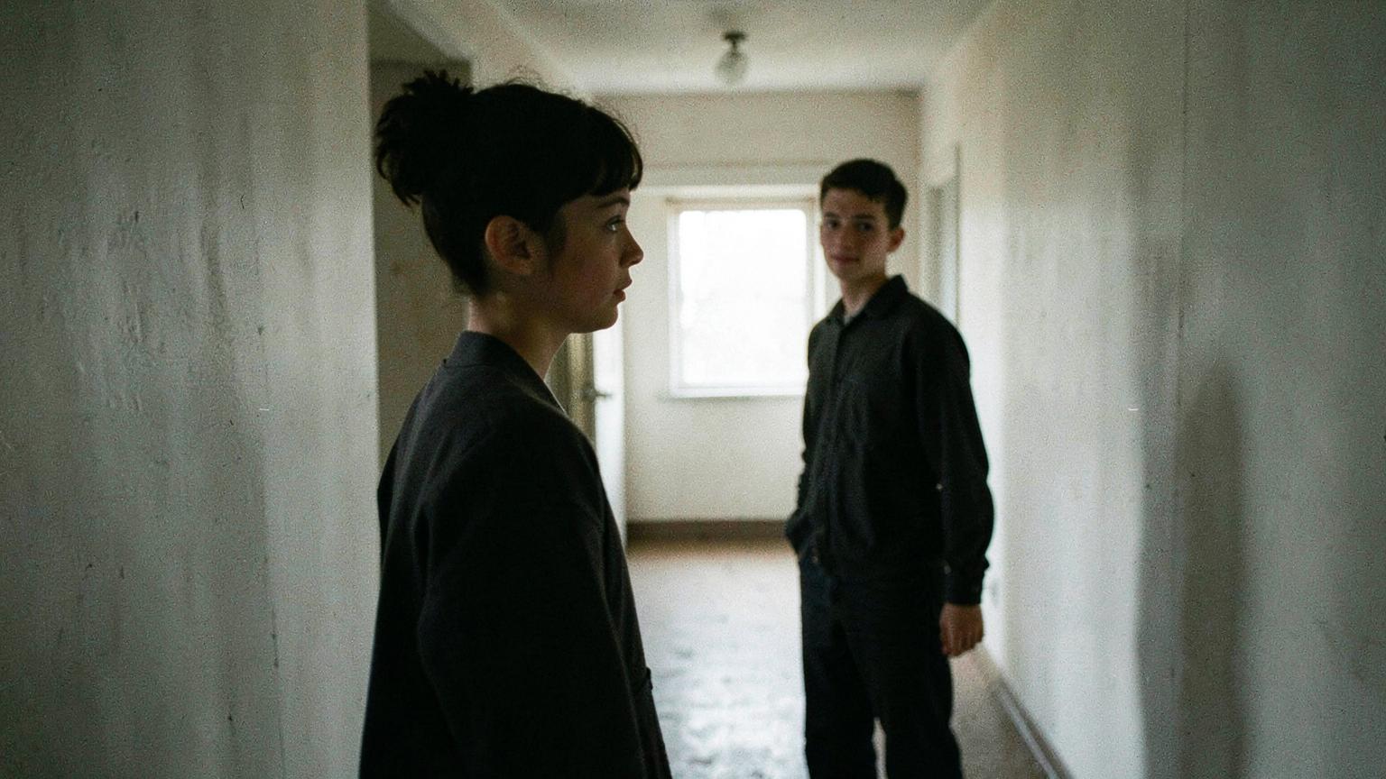 Two young people in a bare hallway, one near the camera in profile, one further away facing them
